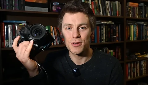 Man holding a mirrorless camera with a prime lens, speaking toward the camera in a home studio setting.