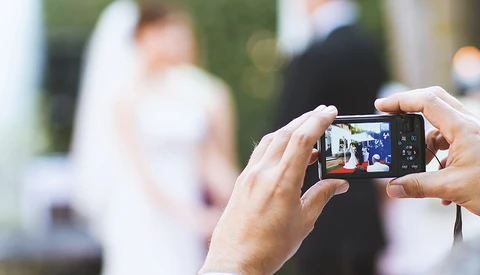 Hands holding a compact digital camera capturing a blurred outdoor event with people in the background.
