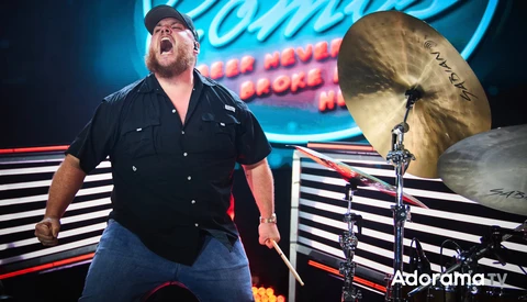 Drummer performing on stage with energetic expression and raised cymbals, vibrant neon backdrop.
