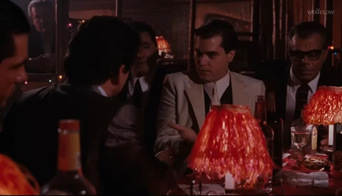 Men in suits gathered around a dinner table with red lampshades and glassware, engaged in conversation during an evening event.