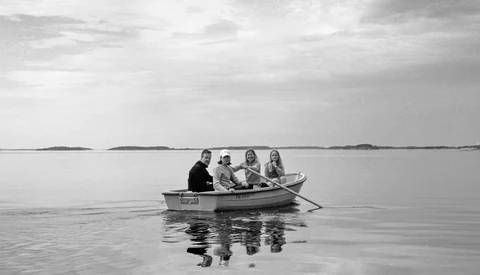 Four people sitting in a small rowboat on calm, glassy water under an overcast sky.