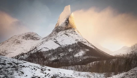 Snow-covered mountain peak with forested slopes under dramatic cloudy sky.
