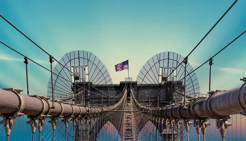 Brooklyn Bridge with symmetrical satellite dishes and American flag, captured with mirrored composition against blue sky.