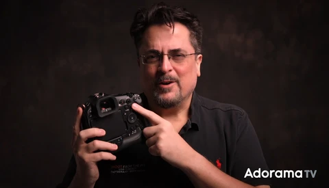 Man with glasses holding a camera controller against a dark background.
