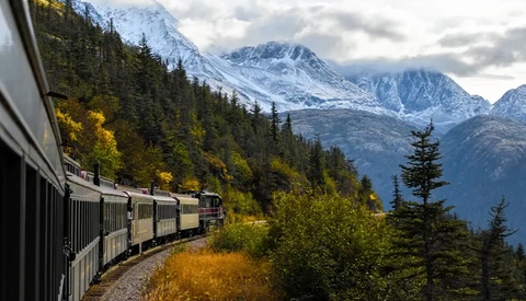 Passenger train curving through forested mountain landscape with snow-capped peaks in background.