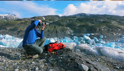 Photographer crouching with camera on tripod, documenting blue glacier icebergs with mountains in background.