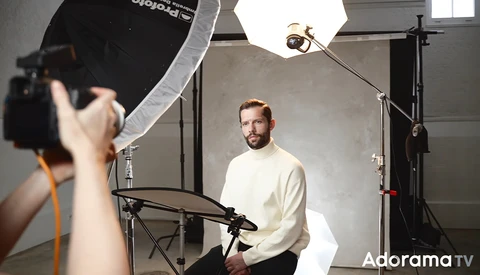 Photographer operating camera while subject sits during studio portrait session with professional lighting setup.