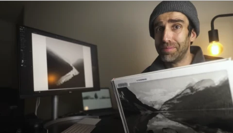 Photographer holding a landscape print while reviewing images on dual monitors in a studio workspace.