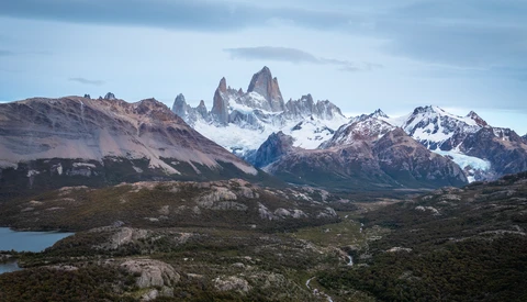 Snow-capped mountain peaks with jagged granite formations rising above a turquoise glacial lake and forested foothills.