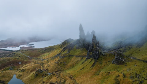 Dramatic mountain peaks shrouded in mist rise above moorland with glacial lakes below.