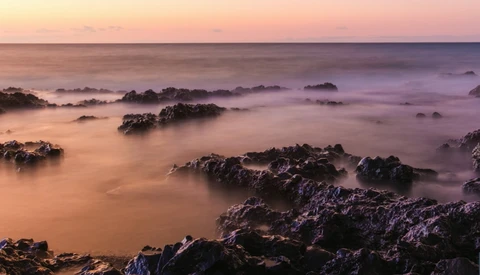Rocky coastline at sunset with smooth water and layered rock formations shrouded in mist.