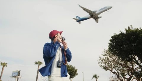 Man in blue jacket and maroon cap watches airplane flying overhead while holding binoculars to his face.
