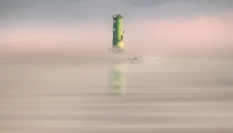 Green and yellow lighthouse emerging from thick fog over calm water.