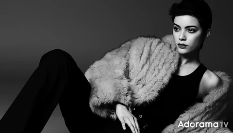 Person in black sleeveless top and fur stole against neutral background, gazing downward.