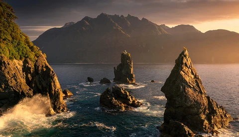 Dramatic rocky sea stacks catching golden sunrise light with misty mountains across the water.