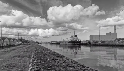 Industrial cargo ship moored alongside a stone quay with residential buildings and modern structures in the background under dramatic cloudy sky.