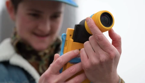 Child holding a yellow and black digital rangefinder device with lens attachment.