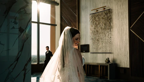 Bride in white lace dress and veil standing in modern architectural interior with groom visible in background.