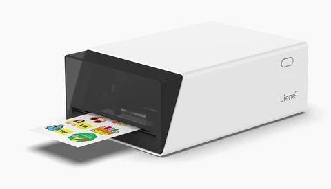 Illustration of a white and black compact photo printer with colorful printed photos emerging from the front tray.
