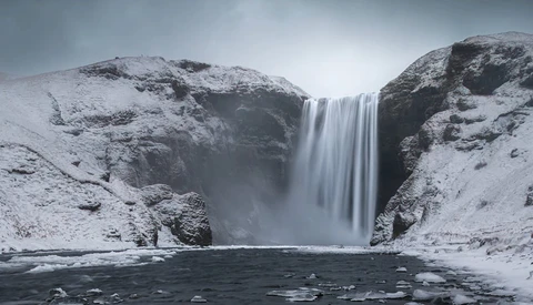 Tall waterfall cascading between snow-covered rocky cliffs into a dark pool of water.