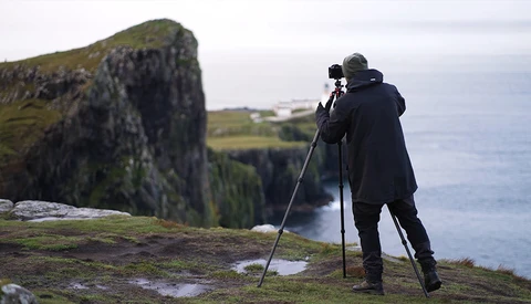 Photographer on tripod at coastal cliff edge, composing seascape with moss-covered rock formation.
