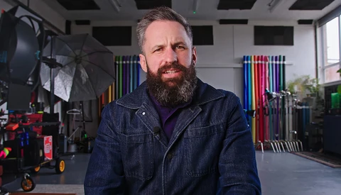 Bearded man in dark denim jacket standing in a professional studio with lighting equipment and colored gels visible in the background.