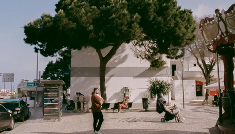 Mediterranean plaza with white building, large tree, and pedestrians in natural daylight.