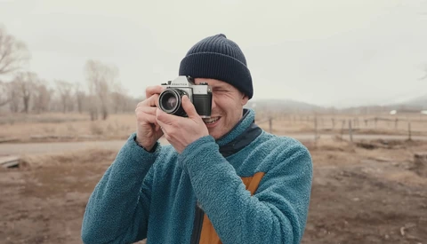 Smiling photographer holding a vintage rangefinder camera at eye level in an open field.
