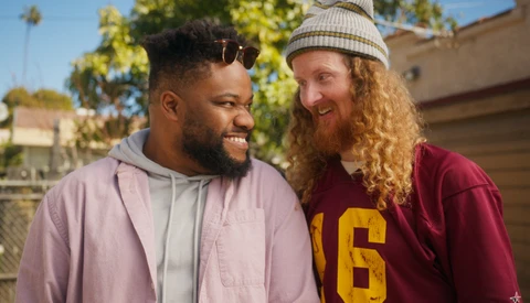 Two smiling men outdoors in casual clothing, one wearing a light pink hoodie and the other in a maroon varsity-style shirt with a beanie.
