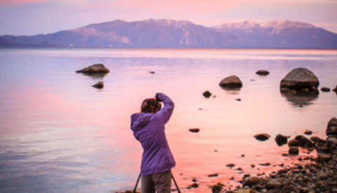 Photographer in purple jacket shooting sunrise landscape with tripod at rocky shoreline.
