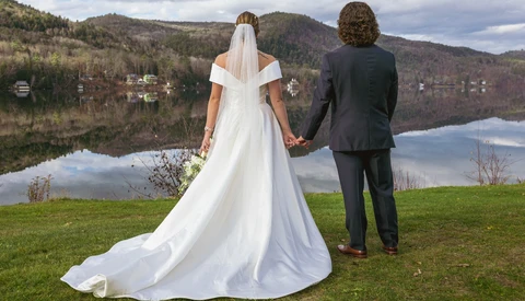 Bride and groom holding hands facing a lake with mountains and forested hills reflected in still water.