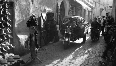 Black and white street scene with vintage car parked among pedestrians in narrow cobblestone alley.
