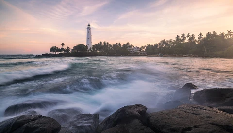 Lighthouse on a peninsula with palm trees, photographed from a rocky beach at golden hour with waves in the foreground.