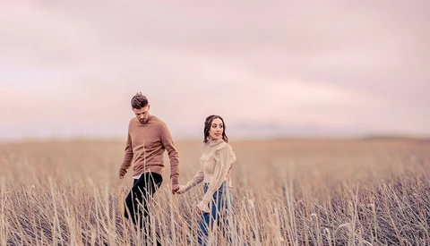 Two people walking through a dried grass field at dusk under a pink-toned sky.