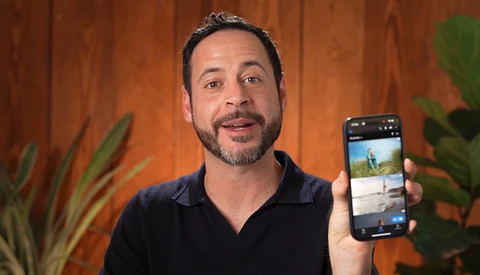 Man in black polo shirt holding smartphone displaying photo app interface against wooden wall with potted plants.