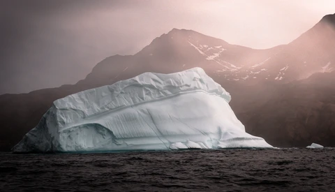 Massive tabular iceberg floating in dark water with snow-capped mountains and soft golden sunlight in the background.