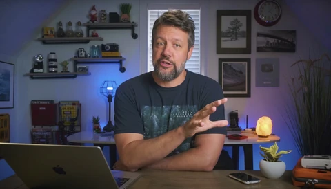 Man in dark t-shirt speaking to camera at desk with laptop and studio lighting setup visible.