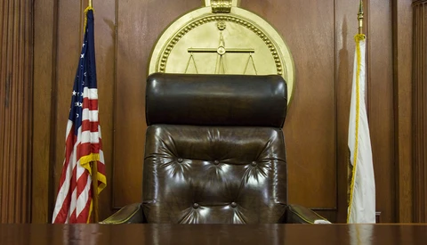 Empty judge's chair in a courtroom with scales of justice symbol behind it.