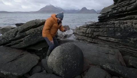 Person in orange jacket exploring rocky coastal landscape with layered basalt formations and mountains across the water.