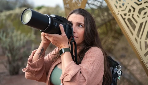 Woman holding vintage film camera up to her face, looking through viewfinder outdoors.