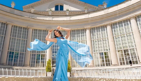 Woman in flowing blue gown with fabric in motion against classical columned building facade.