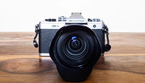 OM System OM-3 rangefinder camera with attached zoom lens on wooden surface.