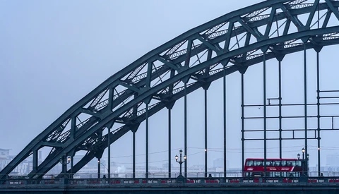 Steel arch bridge structure silhouetted against overcast sky with red double-decker bus below.