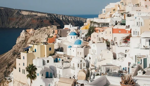 Whitewashed buildings cascading down a clifftop toward turquoise water, with blue-domed church visible among terracotta roofs.