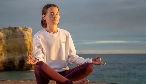 Woman in meditation pose overlooking ocean with rock formations in background.
