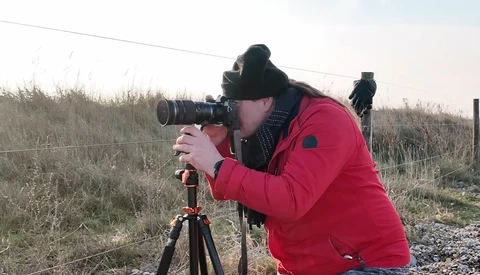 Photographer in red jacket looking through telephoto lens mounted on tripod in rural landscape.