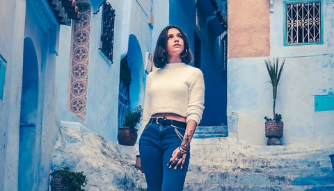 Woman in cream sweater and dark jeans standing against weathered blue and white building wall.