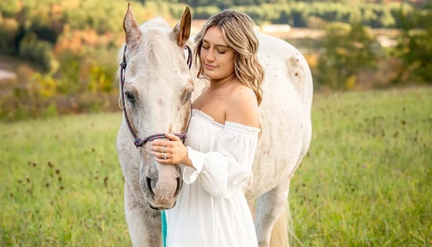 Woman in off-shoulder white dress standing beside a gray horse in a pastoral field.