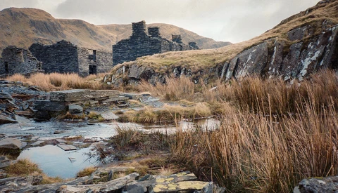 Ruined stone buildings in a moorland valley with a shallow stream and reeds in the foreground.