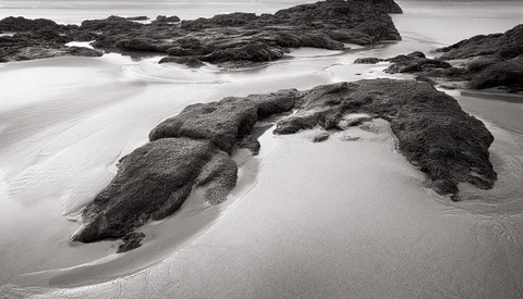 Rocky outcrops emerging from smooth sand on a misty beach with long-exposure water blur.